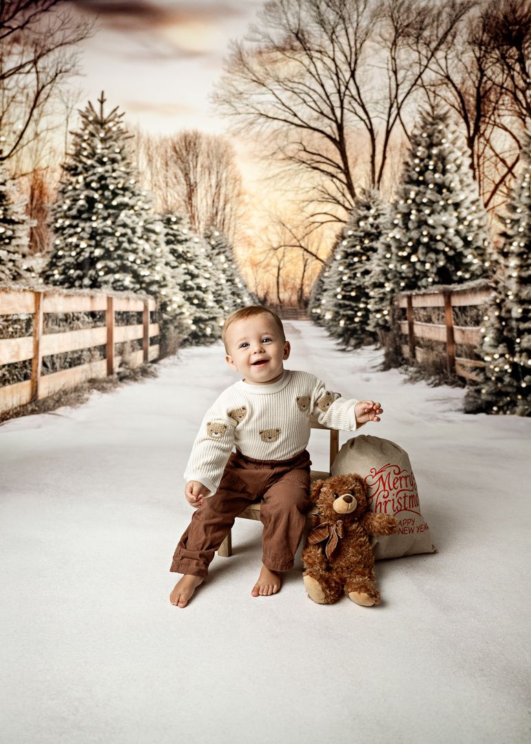 Baby boy in bear sweater sits in snow with teddy bear beside a Christmas sack in a winter wonderland setting.