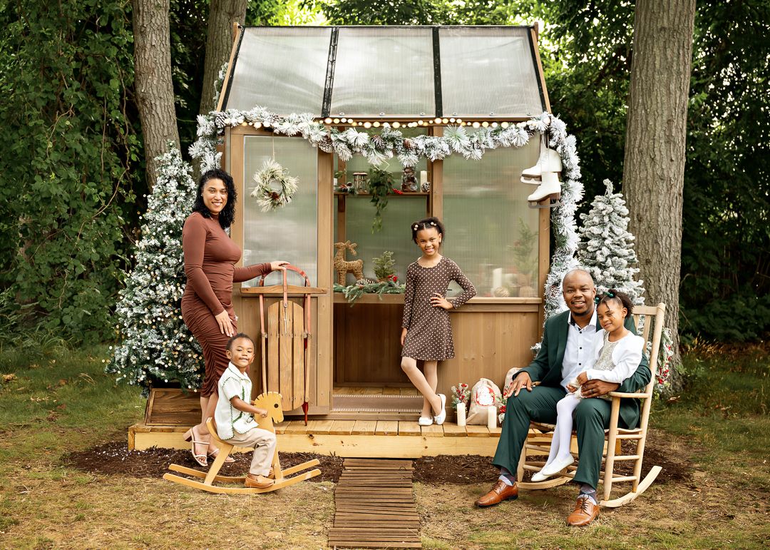 Family posing outdoors in front of a decorated greenhouse with Christmas trees and holiday decor.