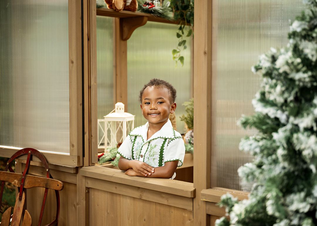 Smiling young child holding a toy dinosaur, standing in a wooden greenhouse doorway decorated for Christmas.