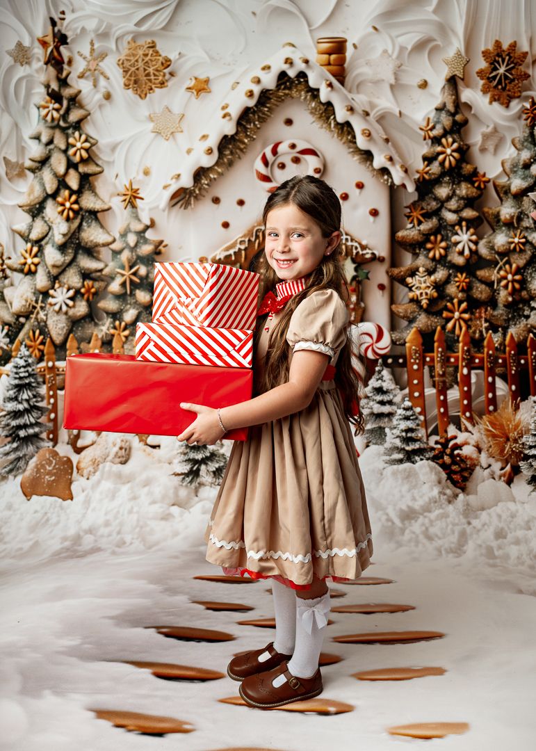 A young girl in a gingerbread-themed dress holds red Christmas gift boxes in a whimsical holiday photo setting.
