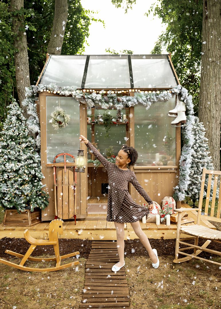 A young girl in a brown polka dot dress dances joyfully outside a Christmas-decorated greenhouse with artificial snow falling.