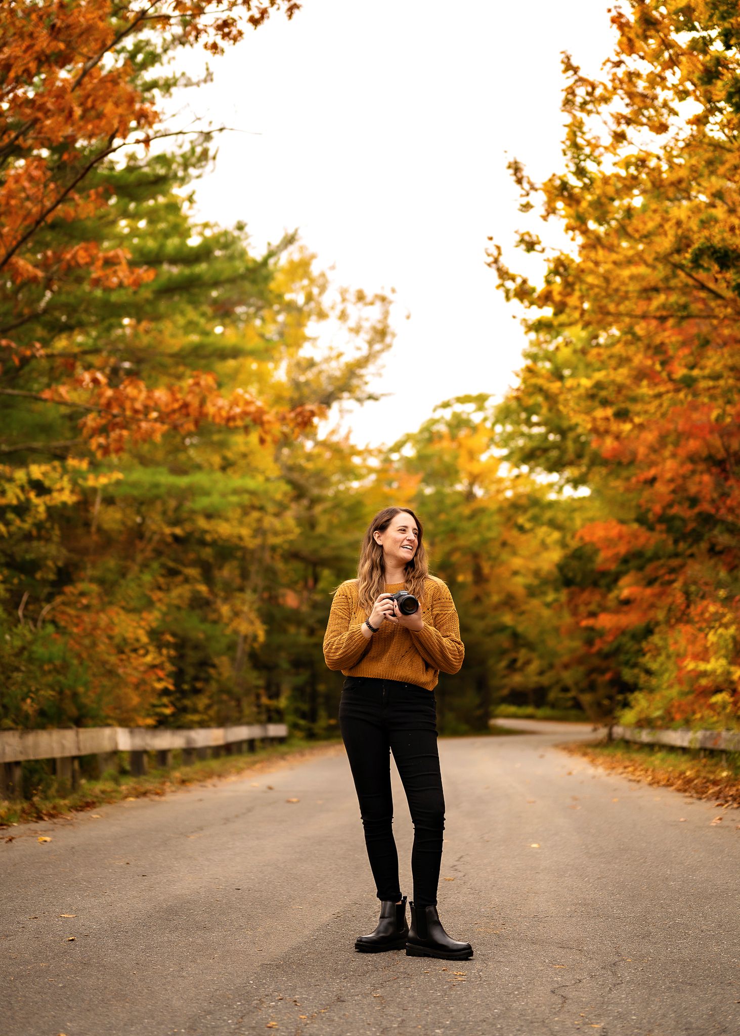 A person in a brown sweater stands on a tree-lined road surrounded by vibrant autumn foliage during golden hour.