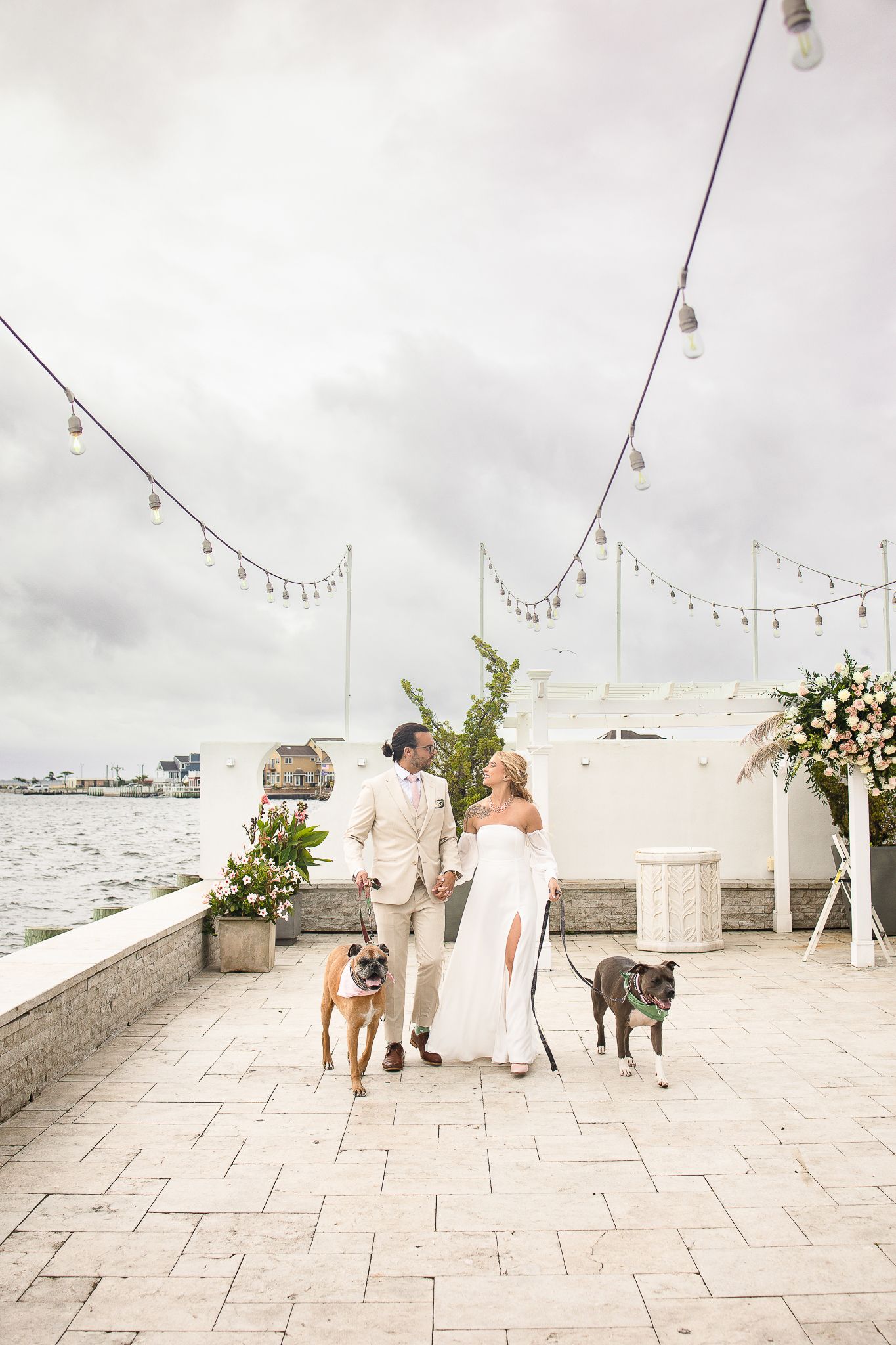 A romantic waterfront ceremony setup with string lights overhead and a couple walking their two dogs on a stone patio.