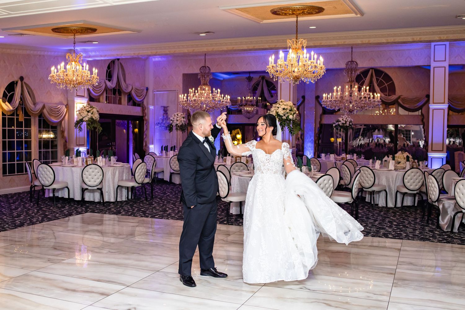Wedding couple performs a choreographed first dance in an elegant ballroom with crystal chandeliers overhead.