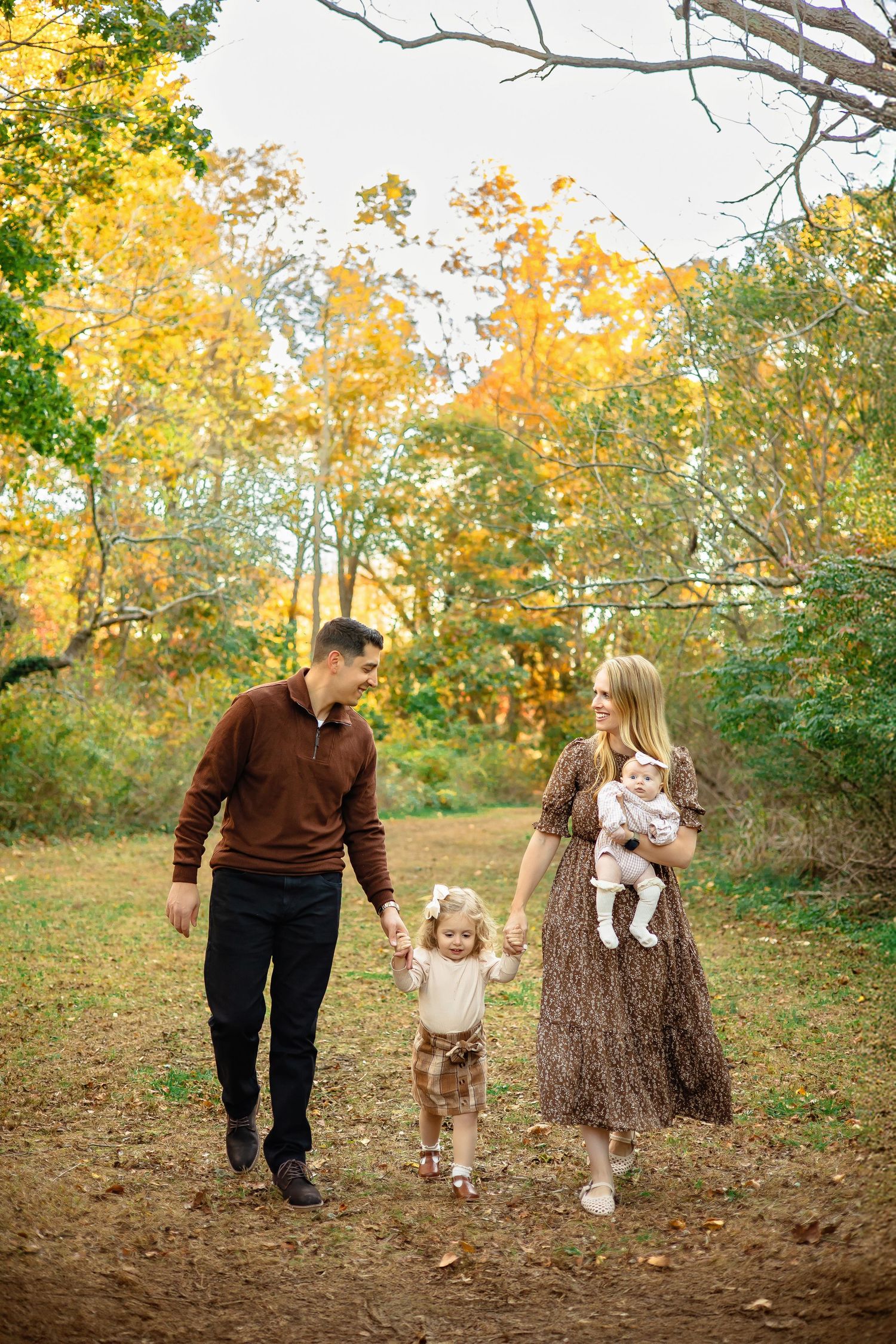 A family walks together on an autumn path surrounded by golden trees and falling leaves in a peaceful woodland setting.