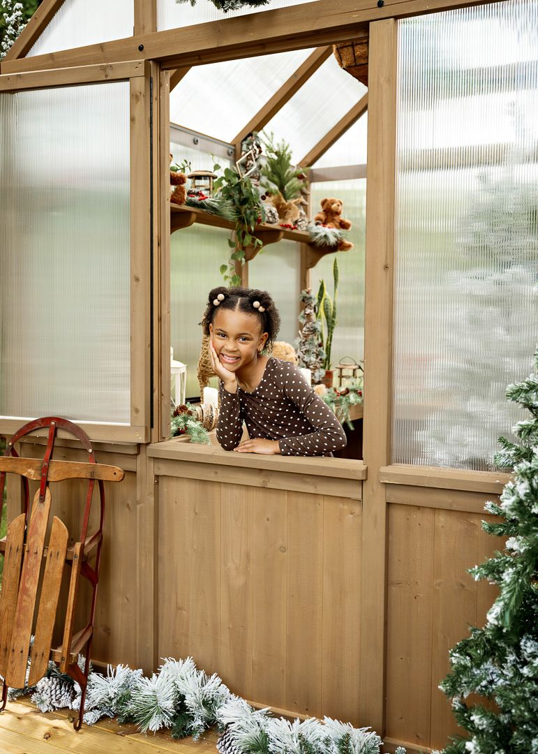 A smiling girl in a brown polka dot dress leans on the windowsill of a Christmas-decorated greenhouse.