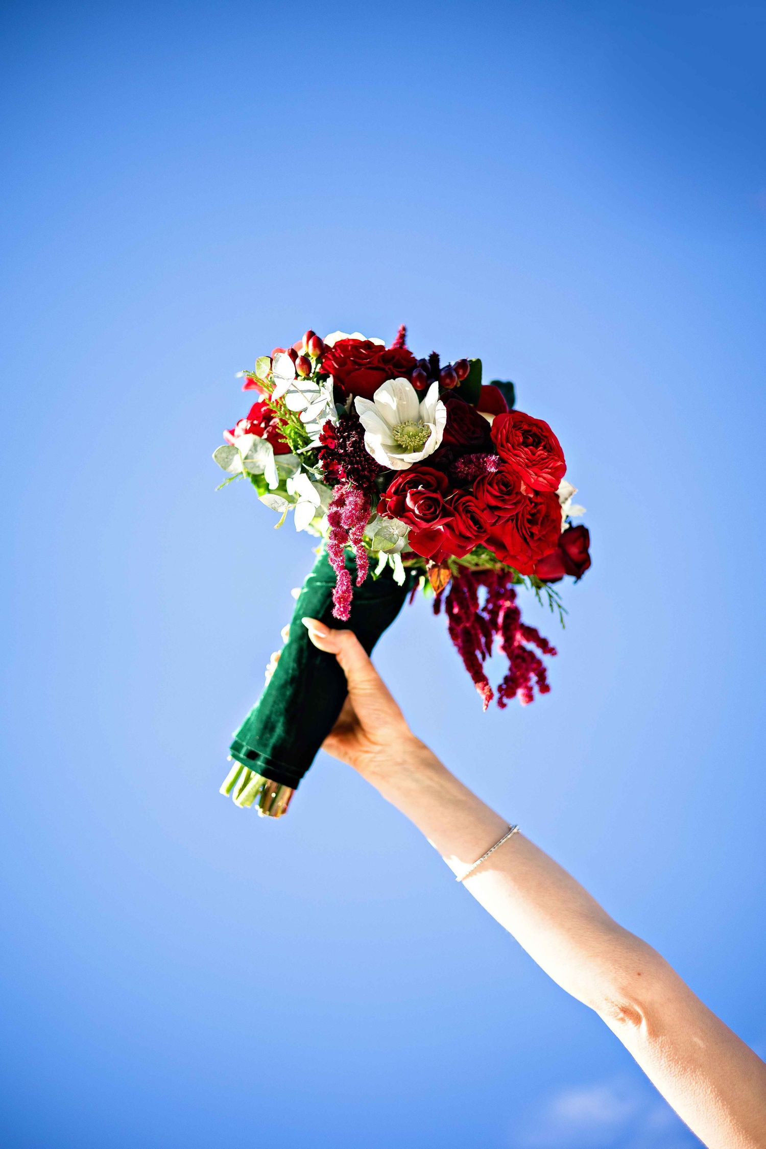 A hand holds up a vibrant red and white bridal bouquet against a bright blue sky background.