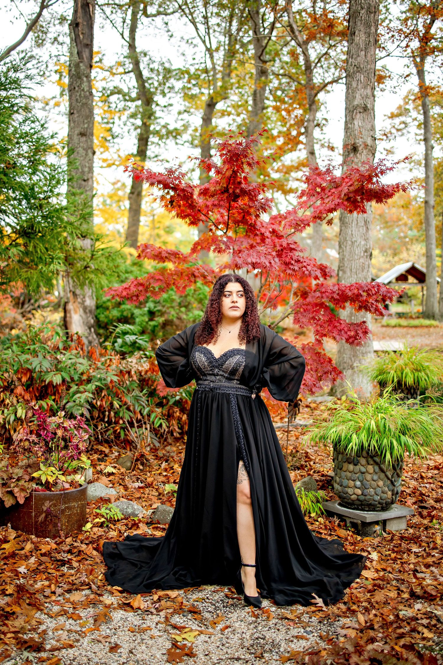 A figure in a flowing black dress poses gracefully against vibrant red autumn foliage in a woodland garden setting.