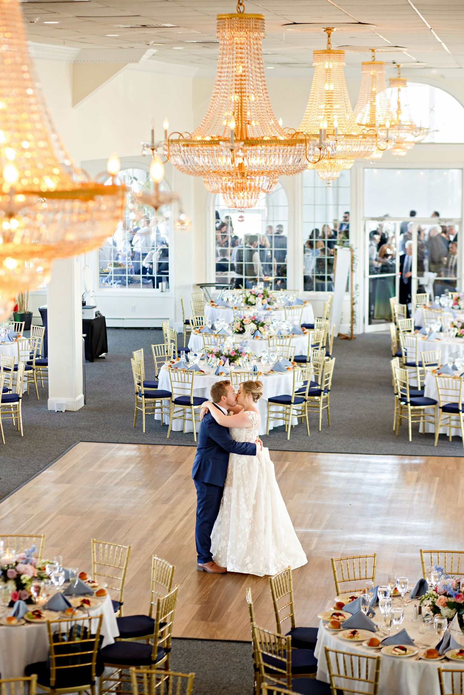 Bride and groom share first dance in elegant wedding reception hall with gold chandeliers and navy blue chiavari chairs.