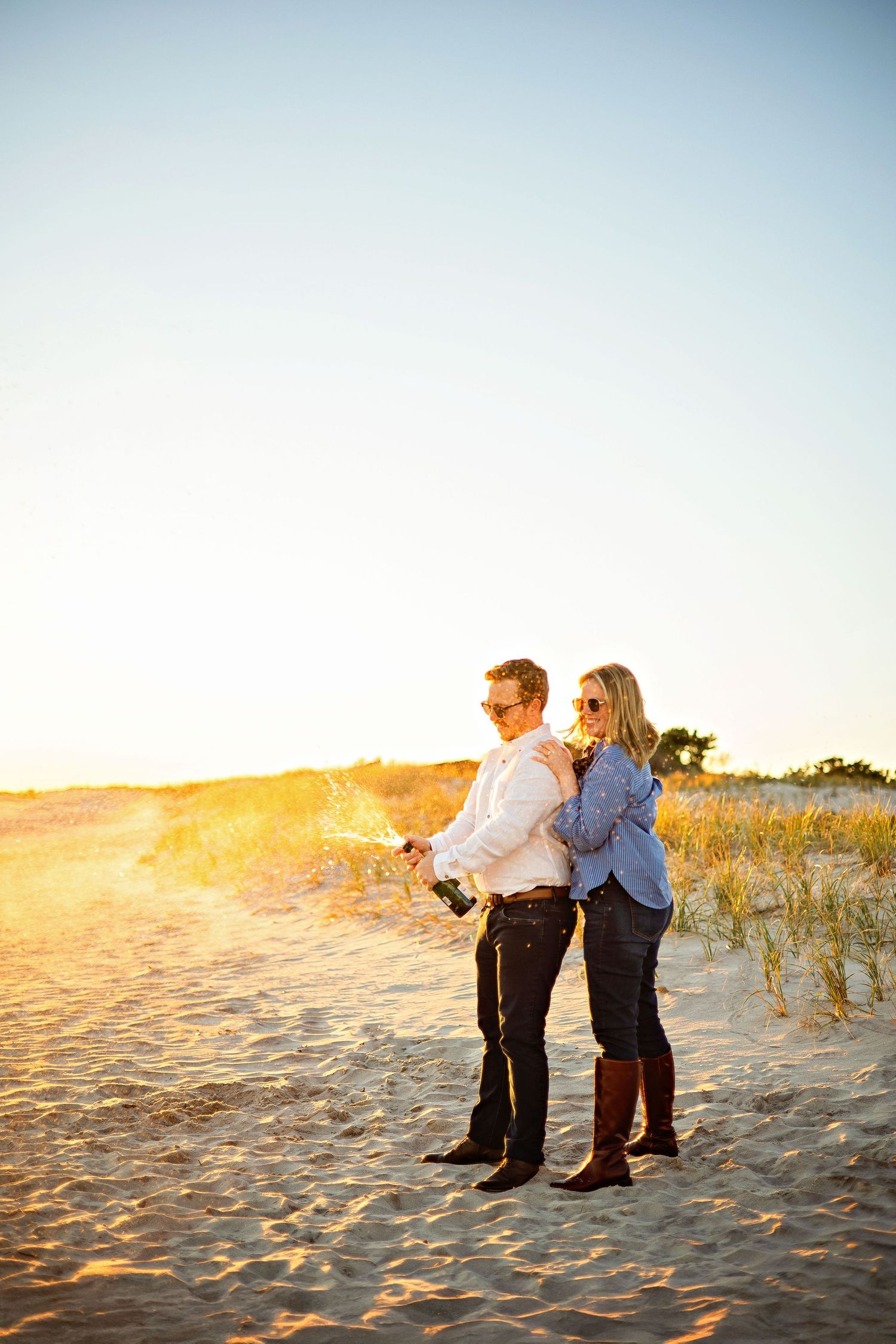 A couple stands together on a sandy beach dune at sunset, creating a romantic silhouette against the golden sky.