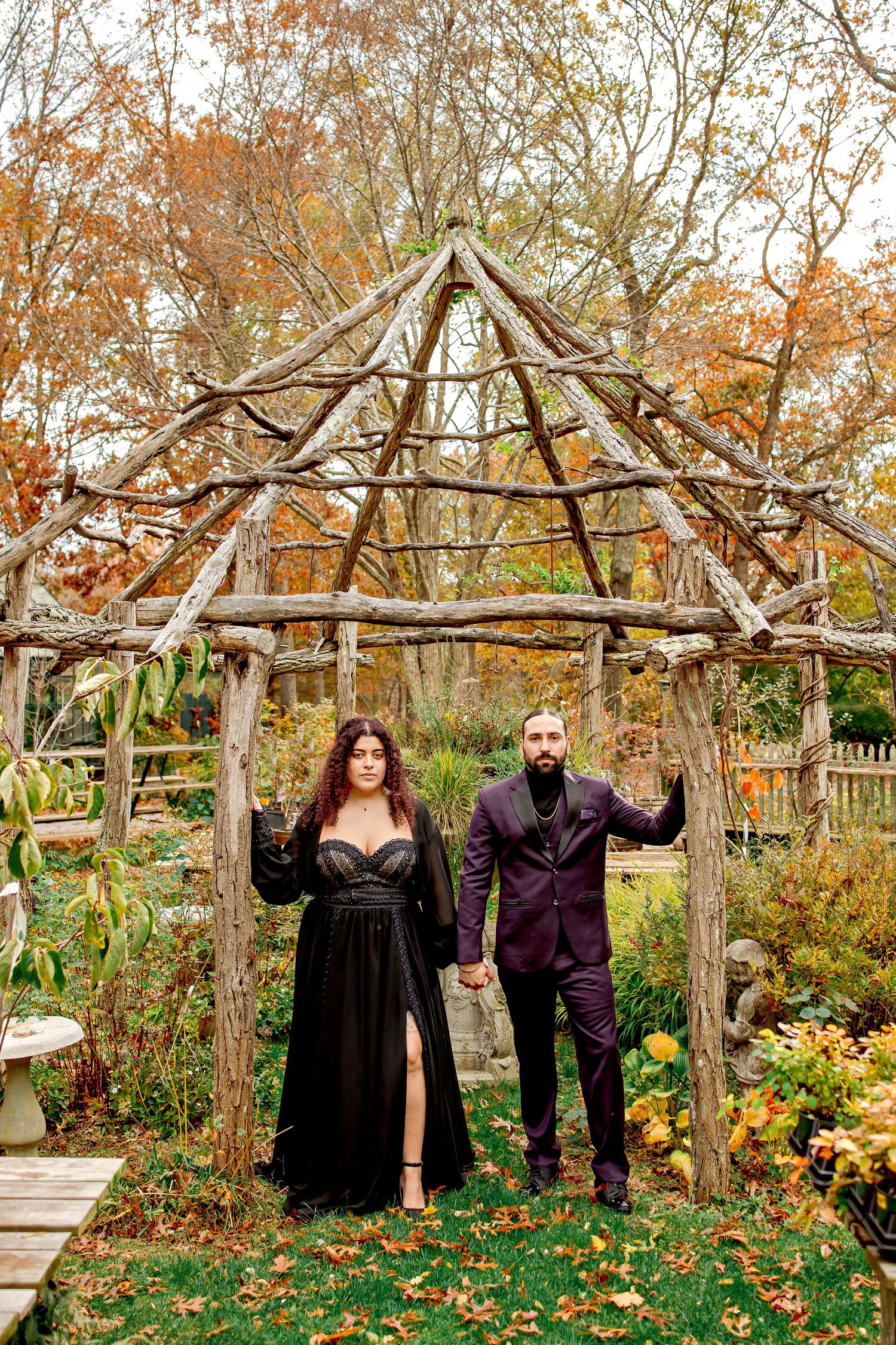 A couple poses under a rustic wooden gazebo surrounded by autumn foliage and garden flowers in a natural setting.
