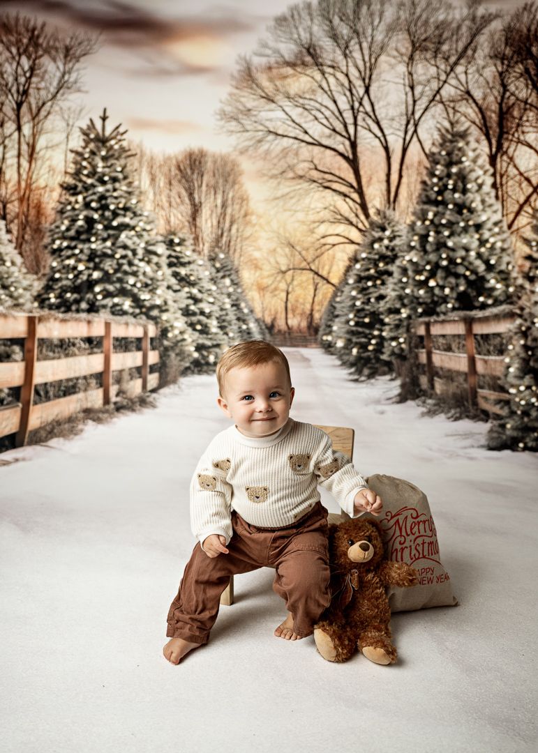 Smiling baby boy in bear sweater sits in snow with teddy bear beside a Christmas tree farm path.
