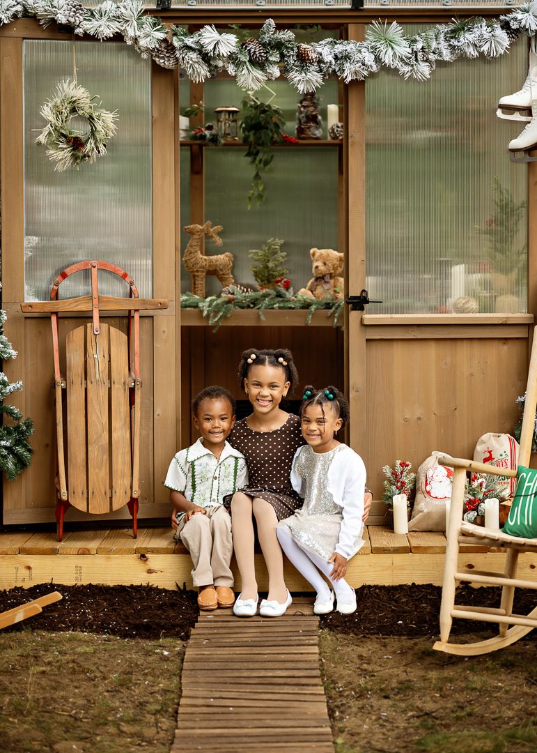 Three children pose together on a decorated wooden porch with Christmas wreaths, a sled, and holiday decor.