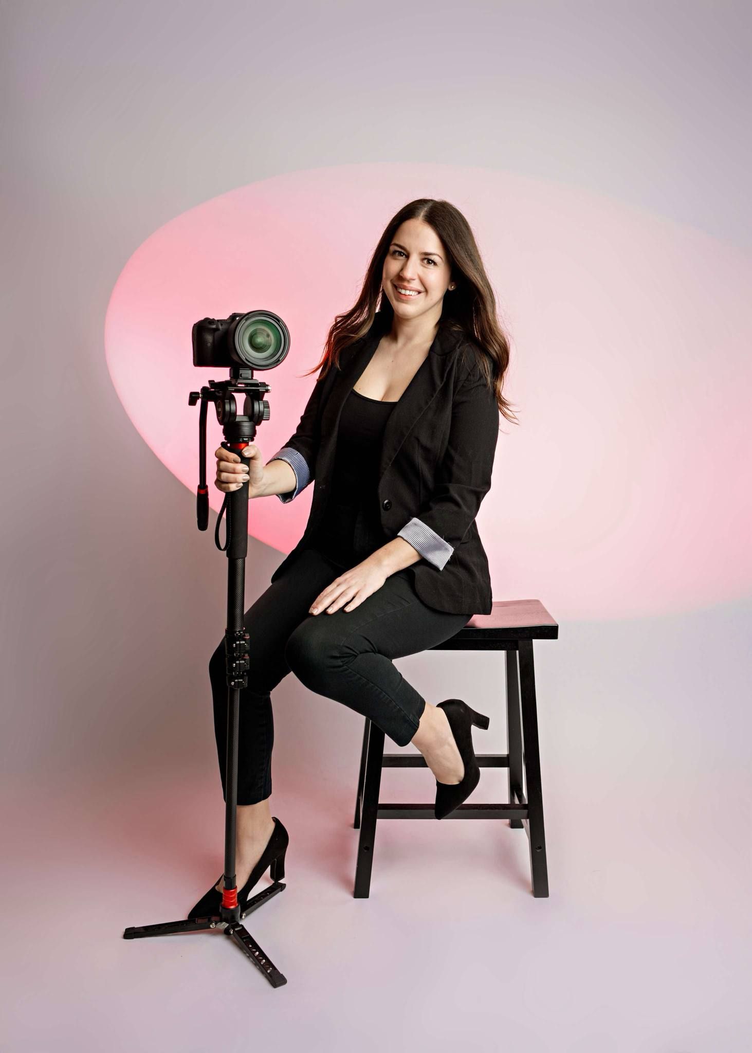 Professional photographer in all black business attire poses with camera equipment against pink backdrop in studio setting.
