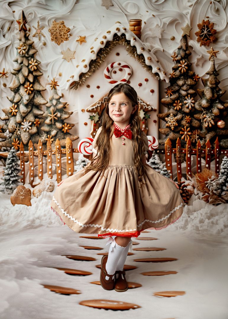 A young girl twirls in a gingerbread costume dress against a magical Christmas gingerbread house backdrop.