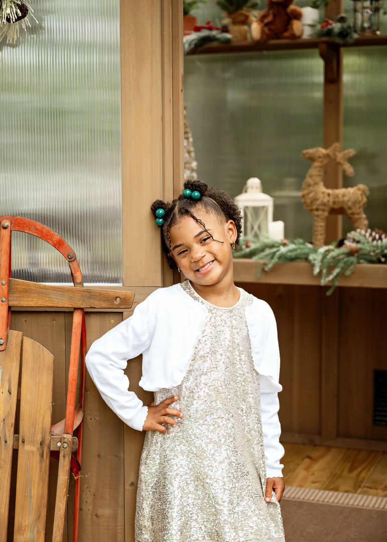 Smiling young girl in sequin dress and white cardigan posing by a festively decorated wooden greenhouse.