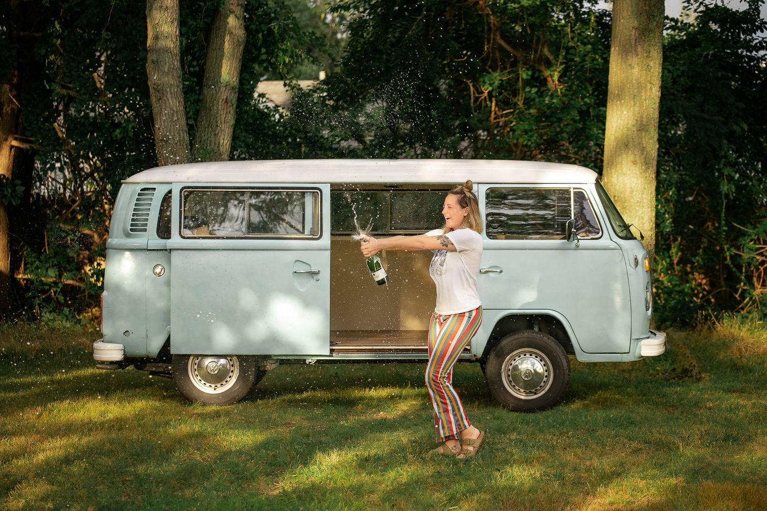 A classic light blue Volkswagen bus parked on grass under trees on a sunny summer day.