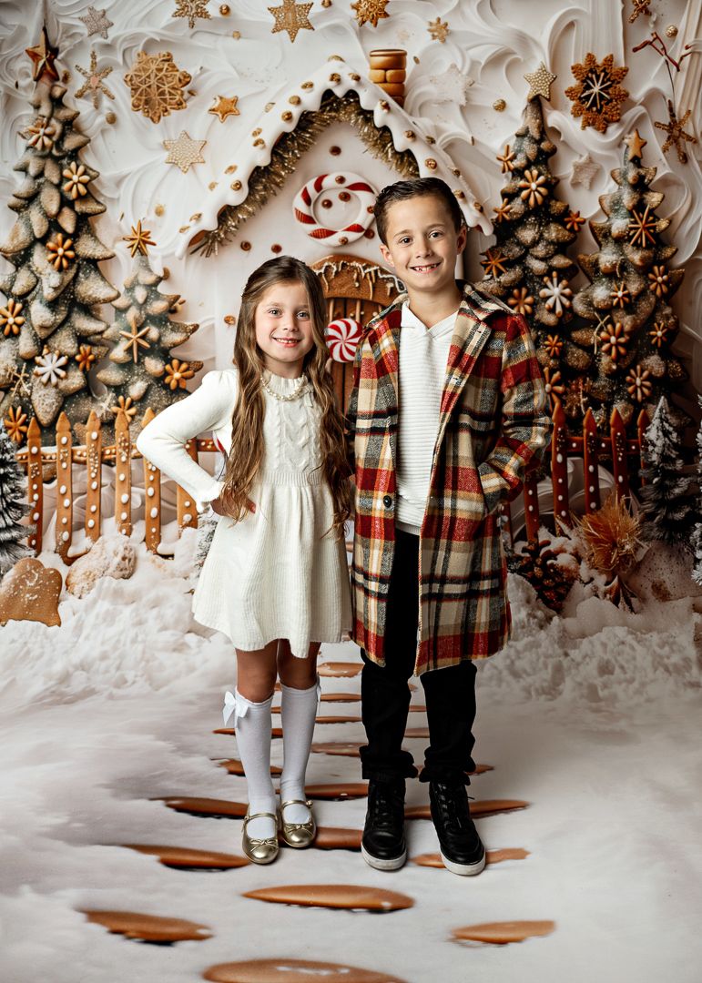 Two children pose before festive gingerbread house backdrop, girl in white dress and boy in plaid coat.