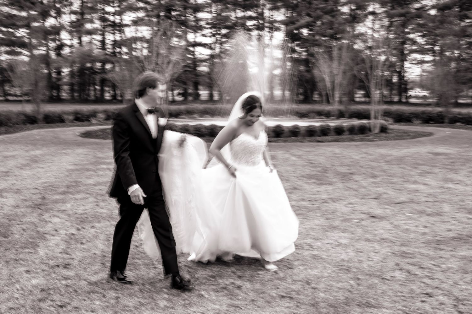 A couple in formal wedding attire walking together on a scenic outdoor path surrounded by pine trees in black and white.