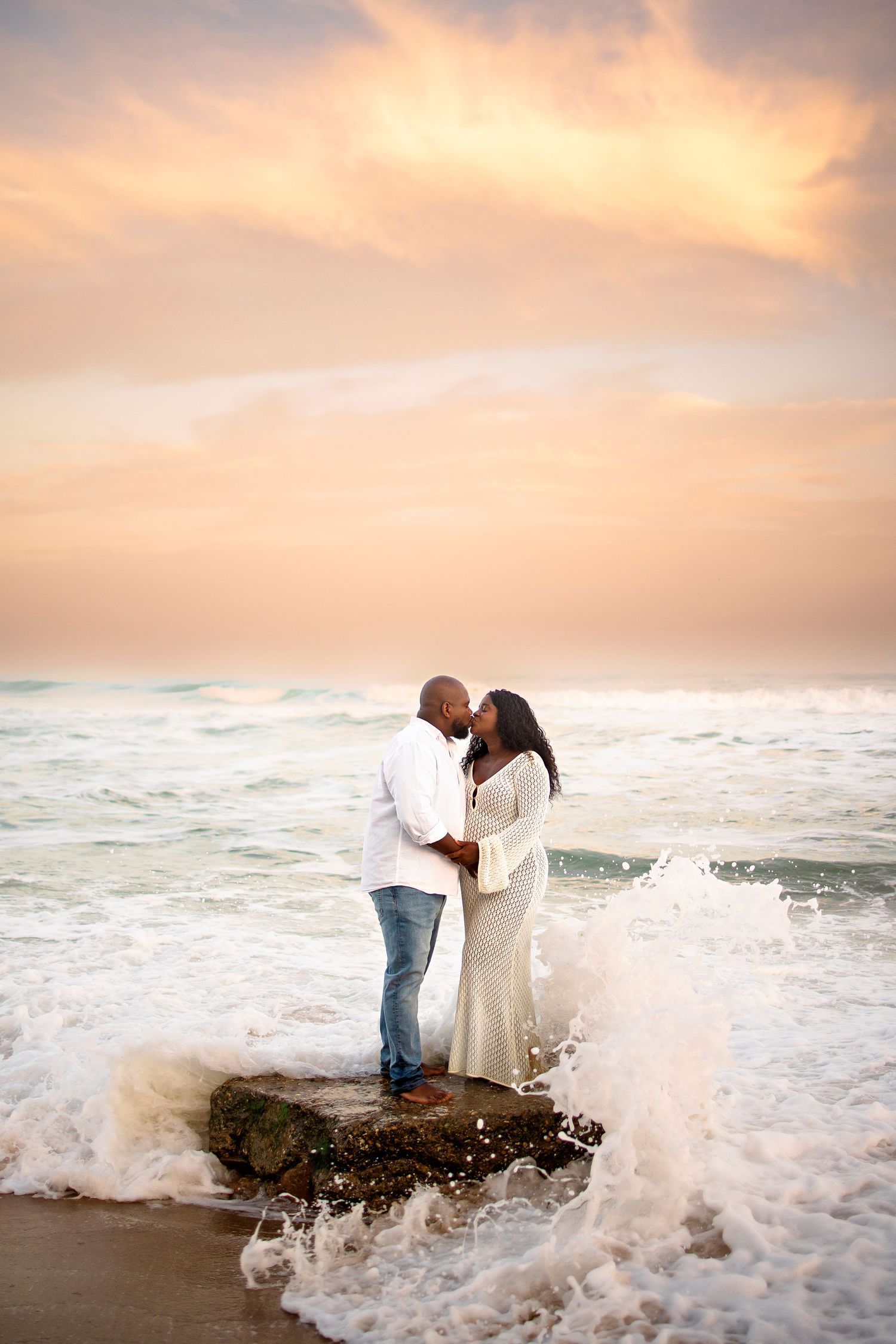 A couple embraces on a rocky beach during sunset as ocean waves crash around them.