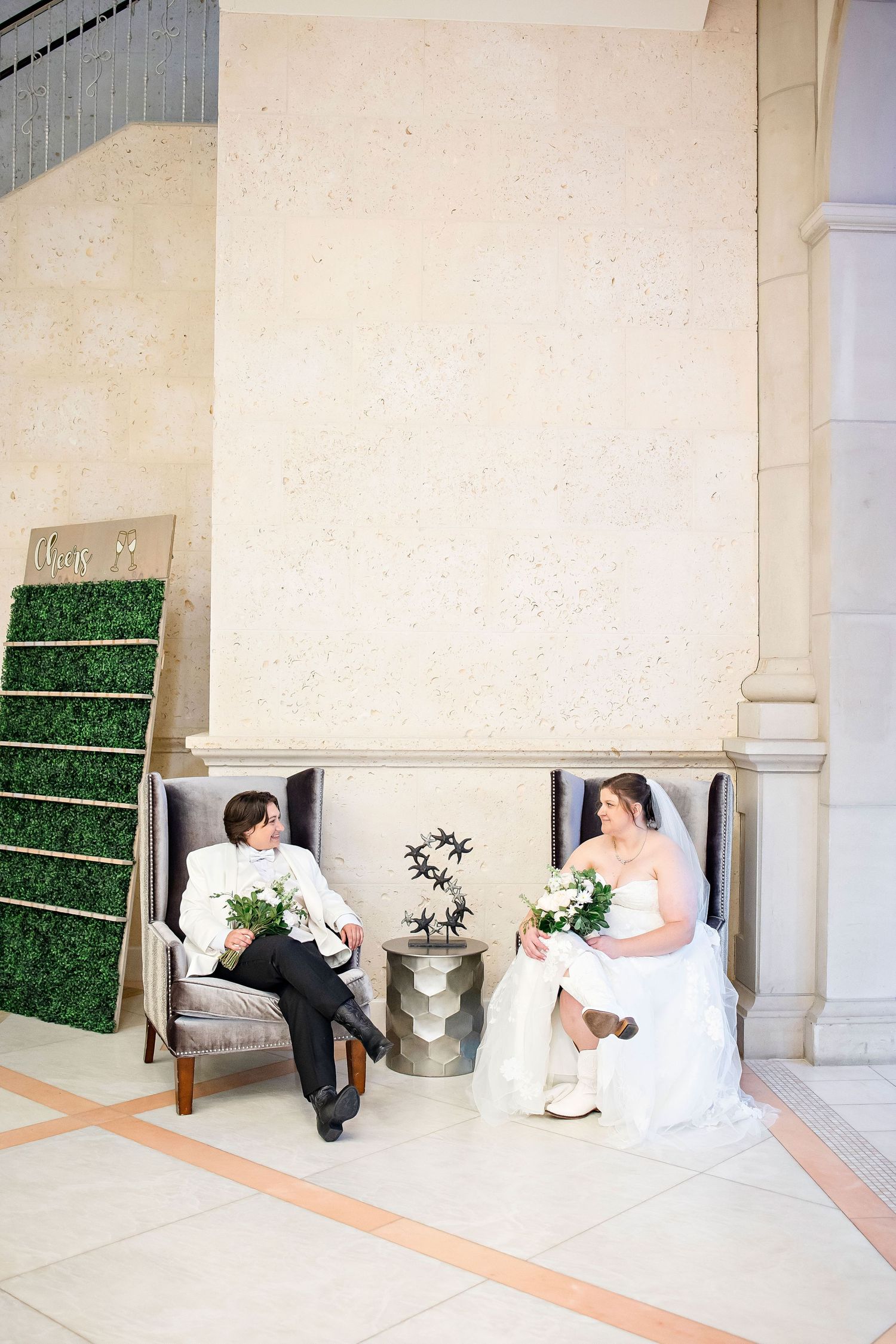 A couple in wedding attire sits apart on elegant chairs with floral bouquets in an ornate white interior space.