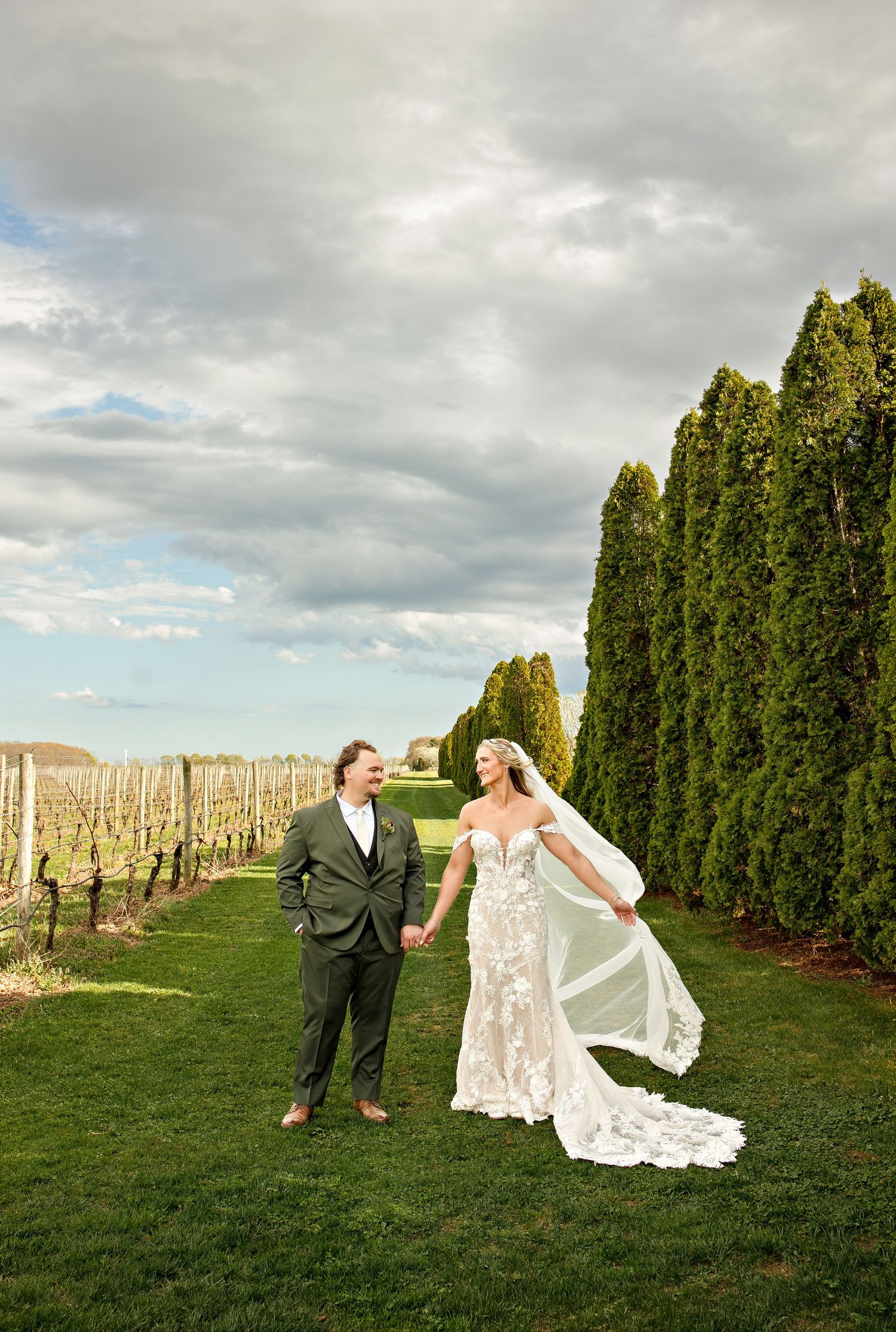 Bride and groom hold hands laughing in a vineyard, her lace gown and veil flowing in the breeze under dramatic clouds.