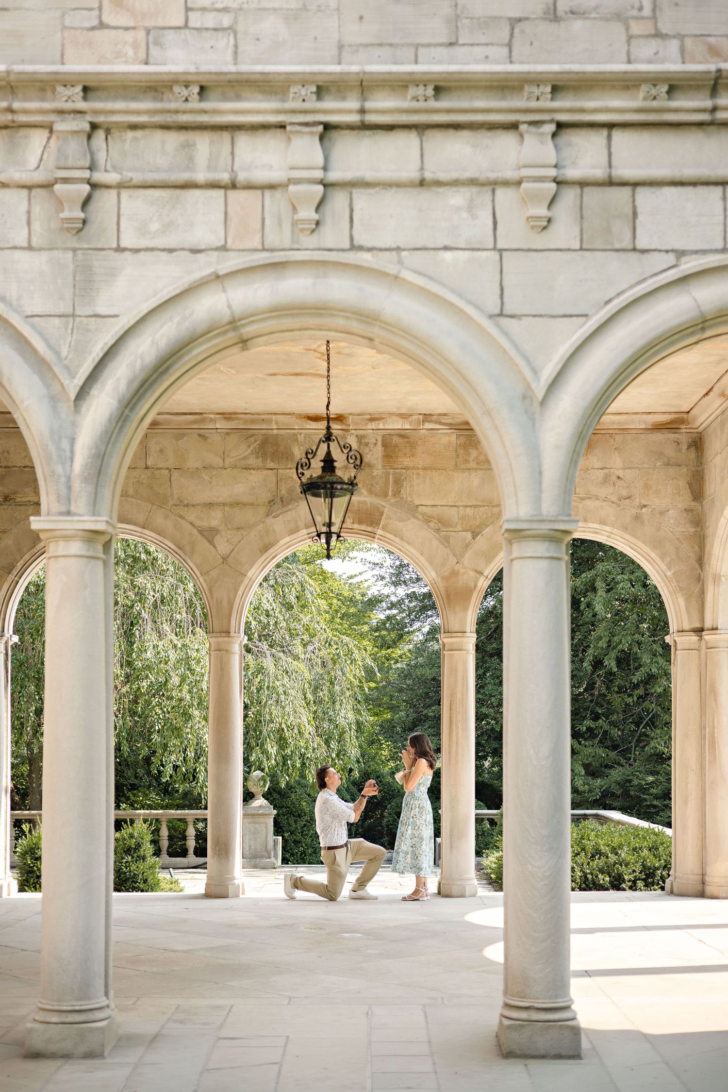 Man proposes to woman under elegant stone arches with hanging lantern in a classical garden setting.