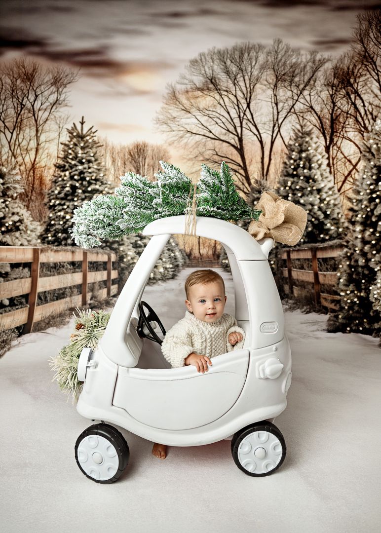 Baby in white sweater sits in toy car with Christmas tree on top, surrounded by snowy winter backdrop.