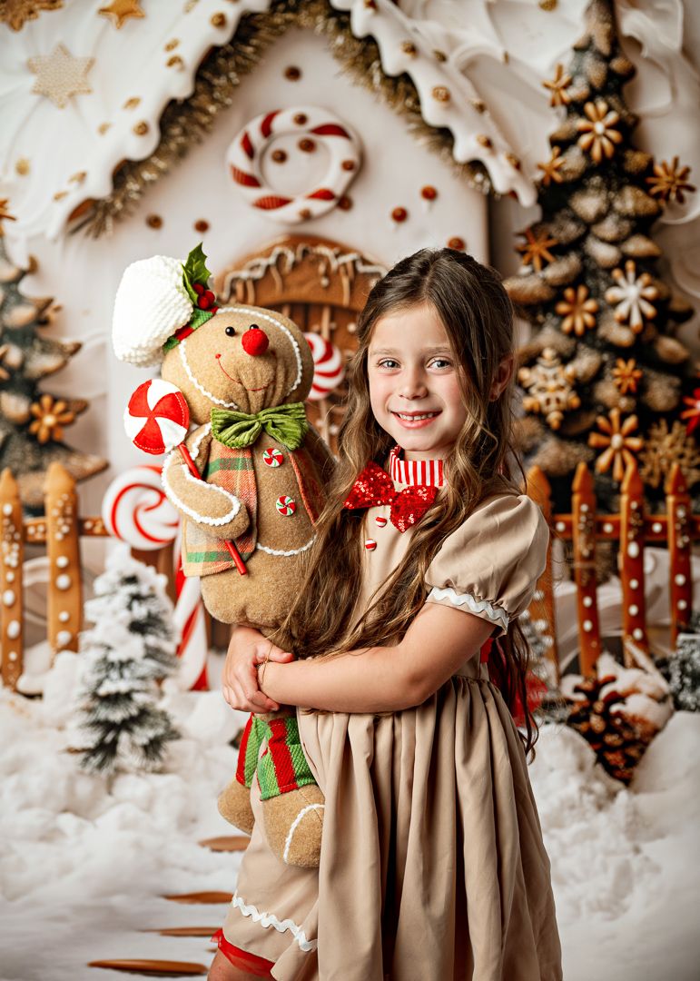 A smiling girl in a beige dress holds a gingerbread man plush toy in a festive Christmas holiday photo backdrop.