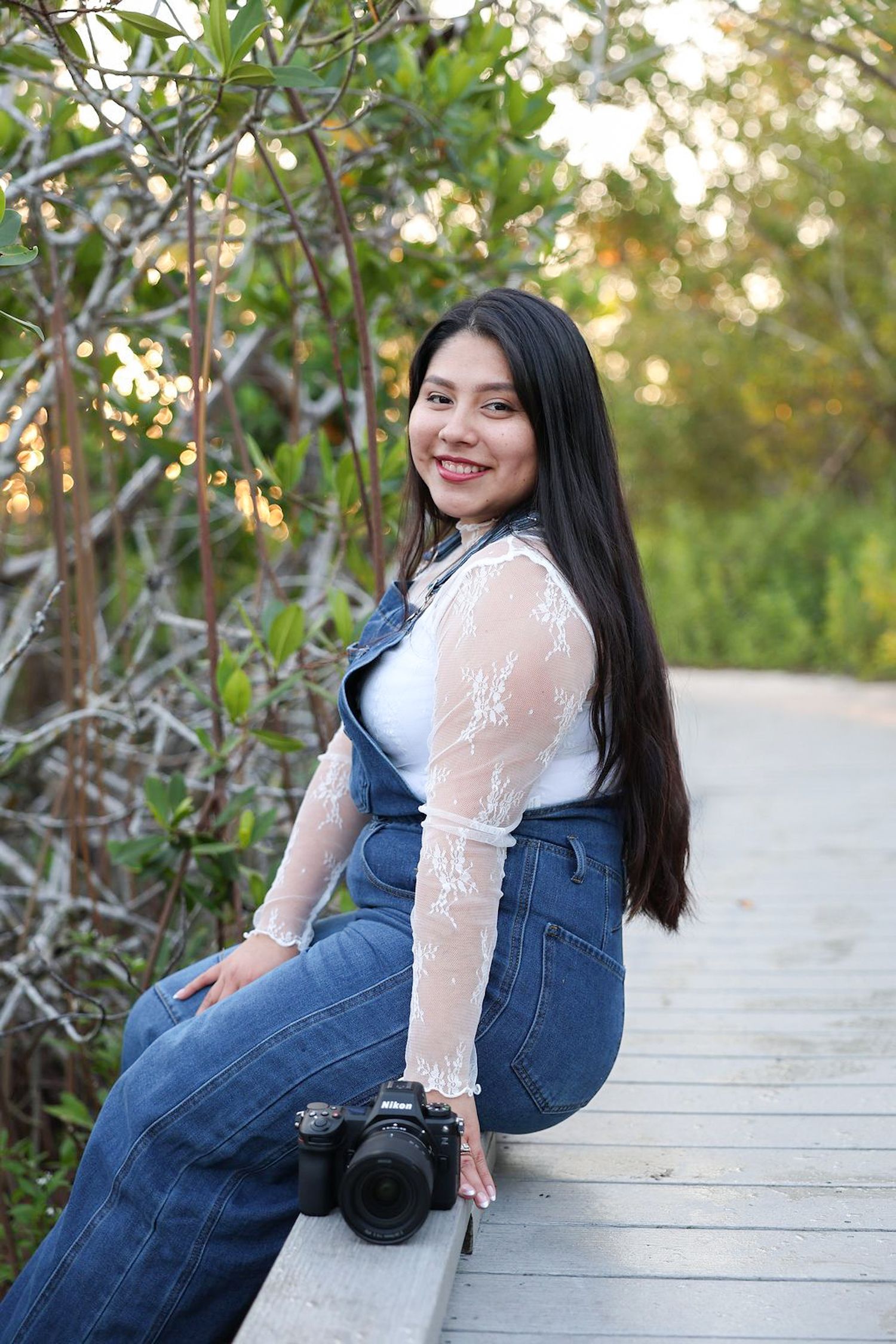 Young woman with long dark hair sits on a wooden boardwalk holding a camera, wearing jeans and lace top.