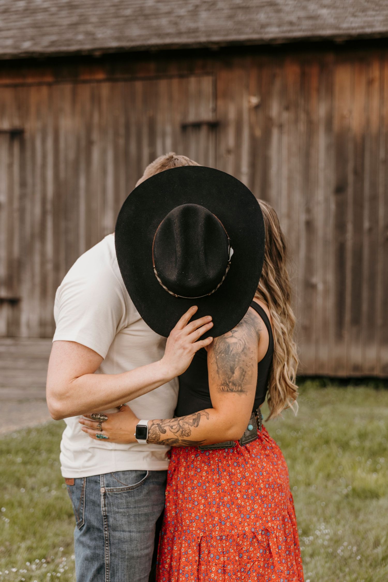 Romantic couple embraces near wooden barn, with black hat and floral skirt details.