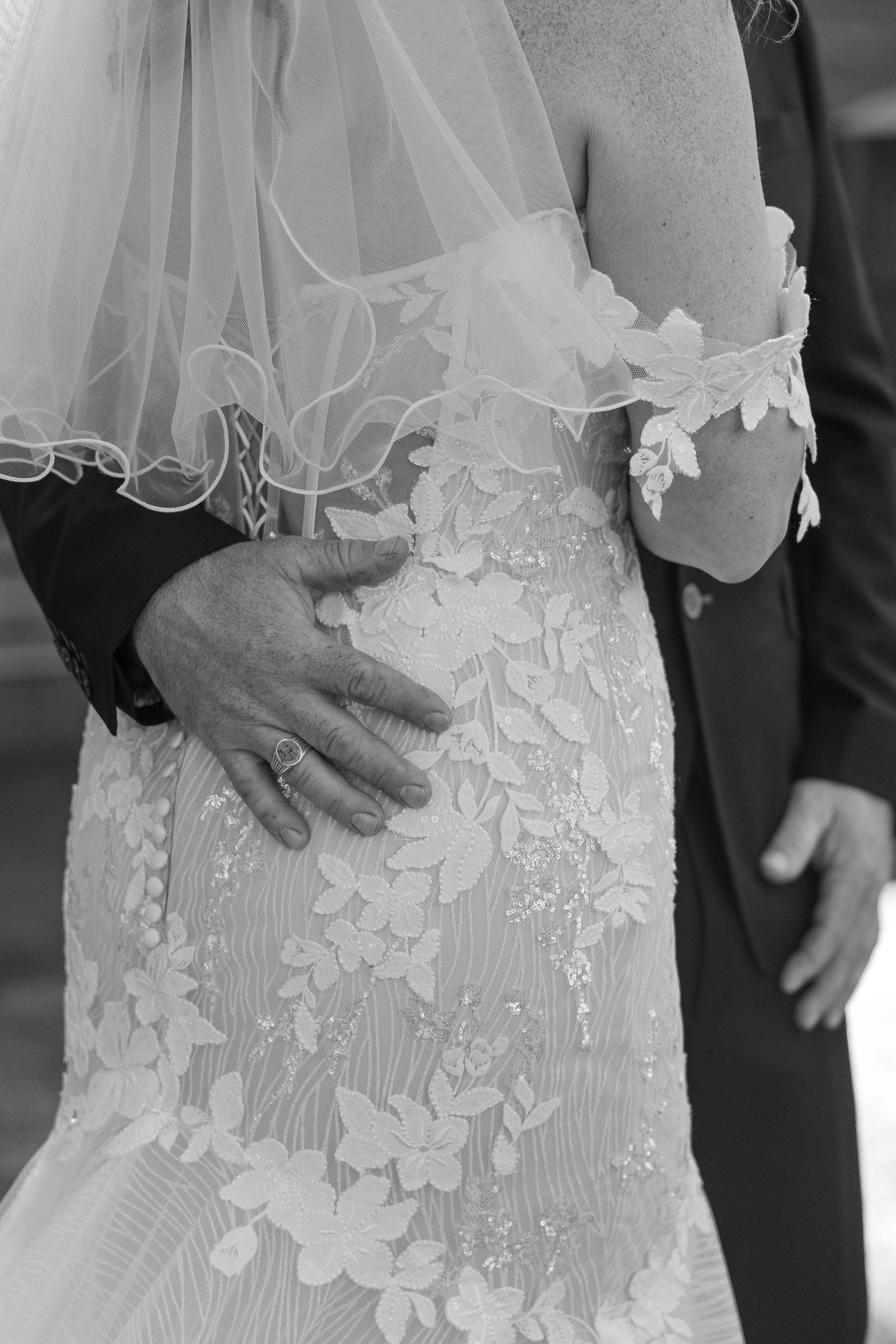 Close-up black and white photo of intricate lace details on a wedding dress with hands embracing the waist.