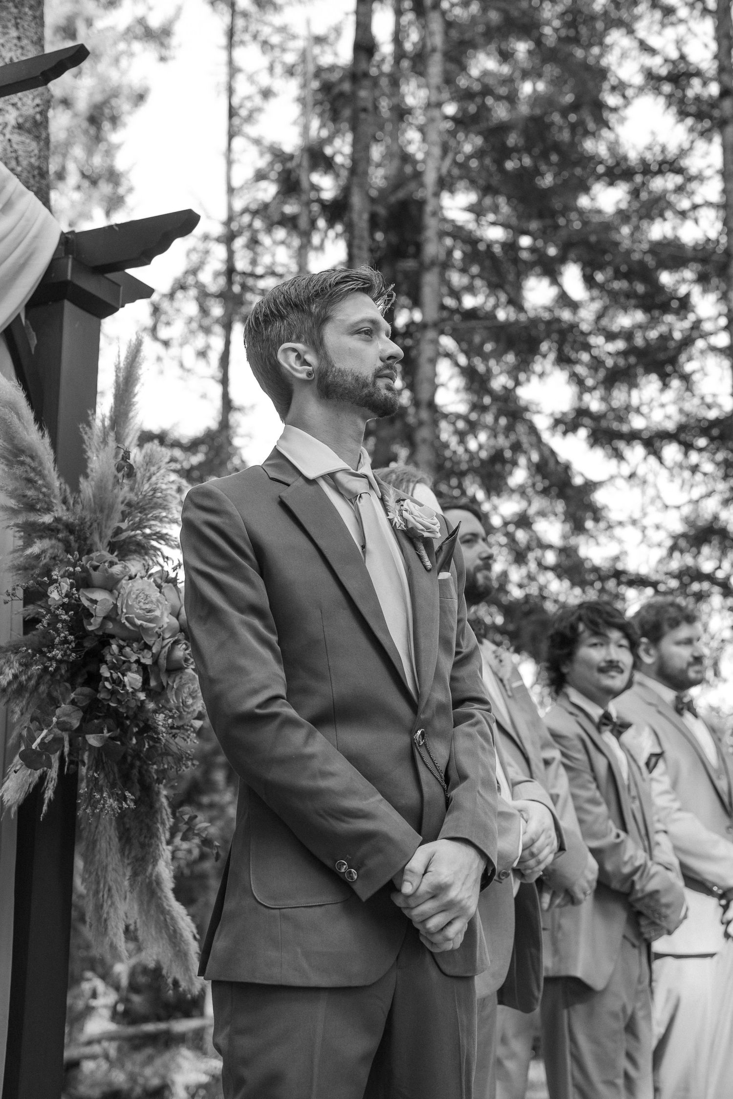 Wedding party members stand in a line during an outdoor ceremony among pine trees.