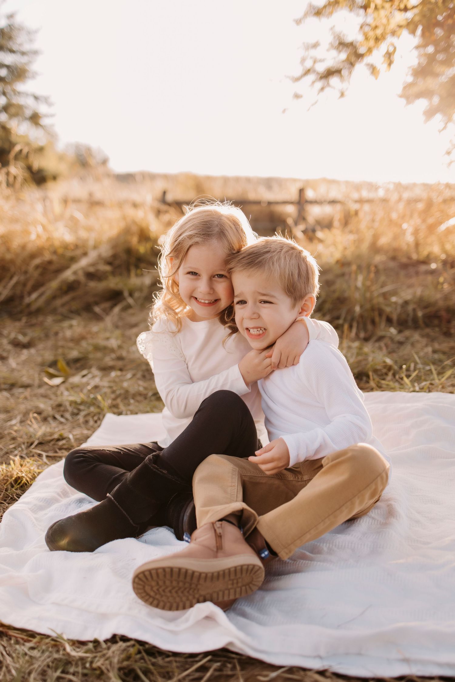 Two children laughing and hugging while sitting on a white blanket in a sunny field.