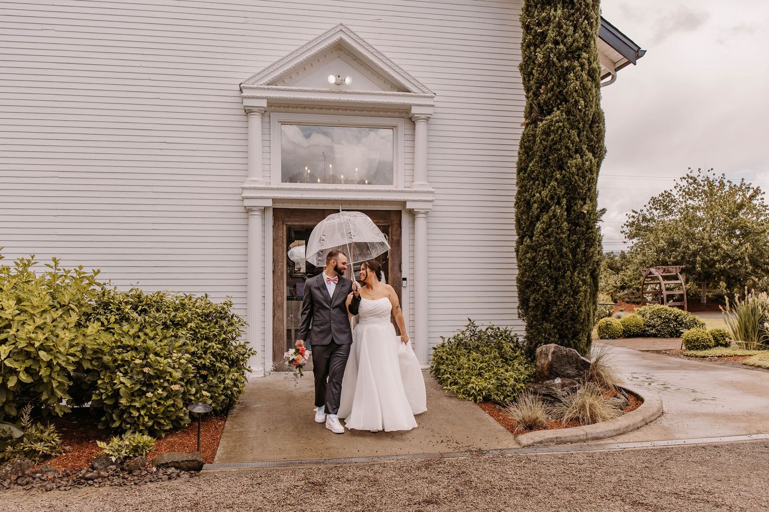 Bride and groom share umbrella outside white chapel on rainy wedding day.