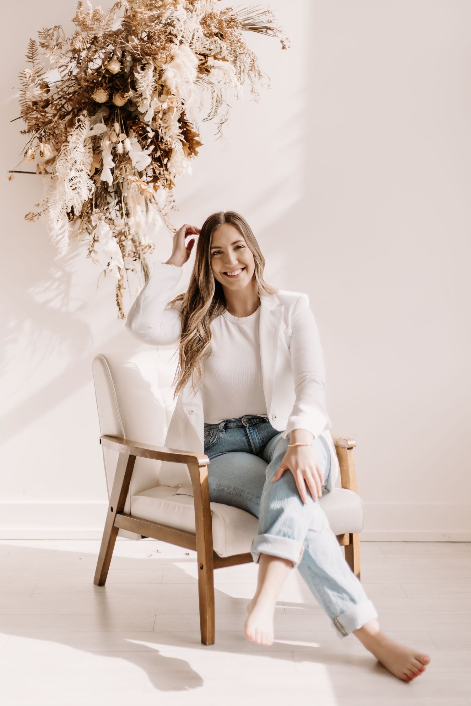 Person in white shirt and jeans sitting in wooden chair with dried floral arrangement overhead.