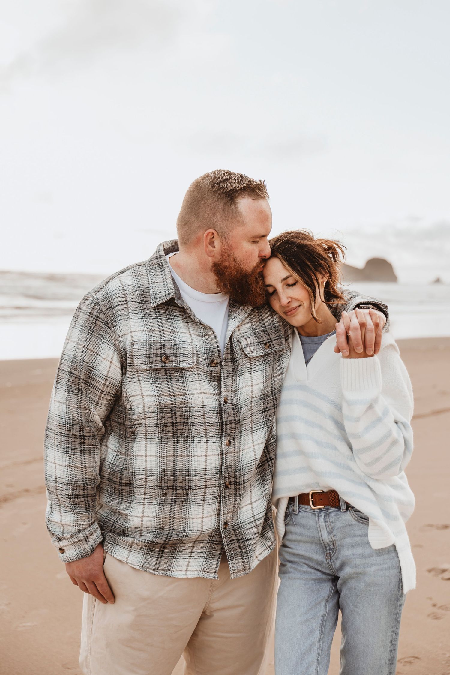 Romantic couple embraces on a sandy beach at sunset wearing casual attire.