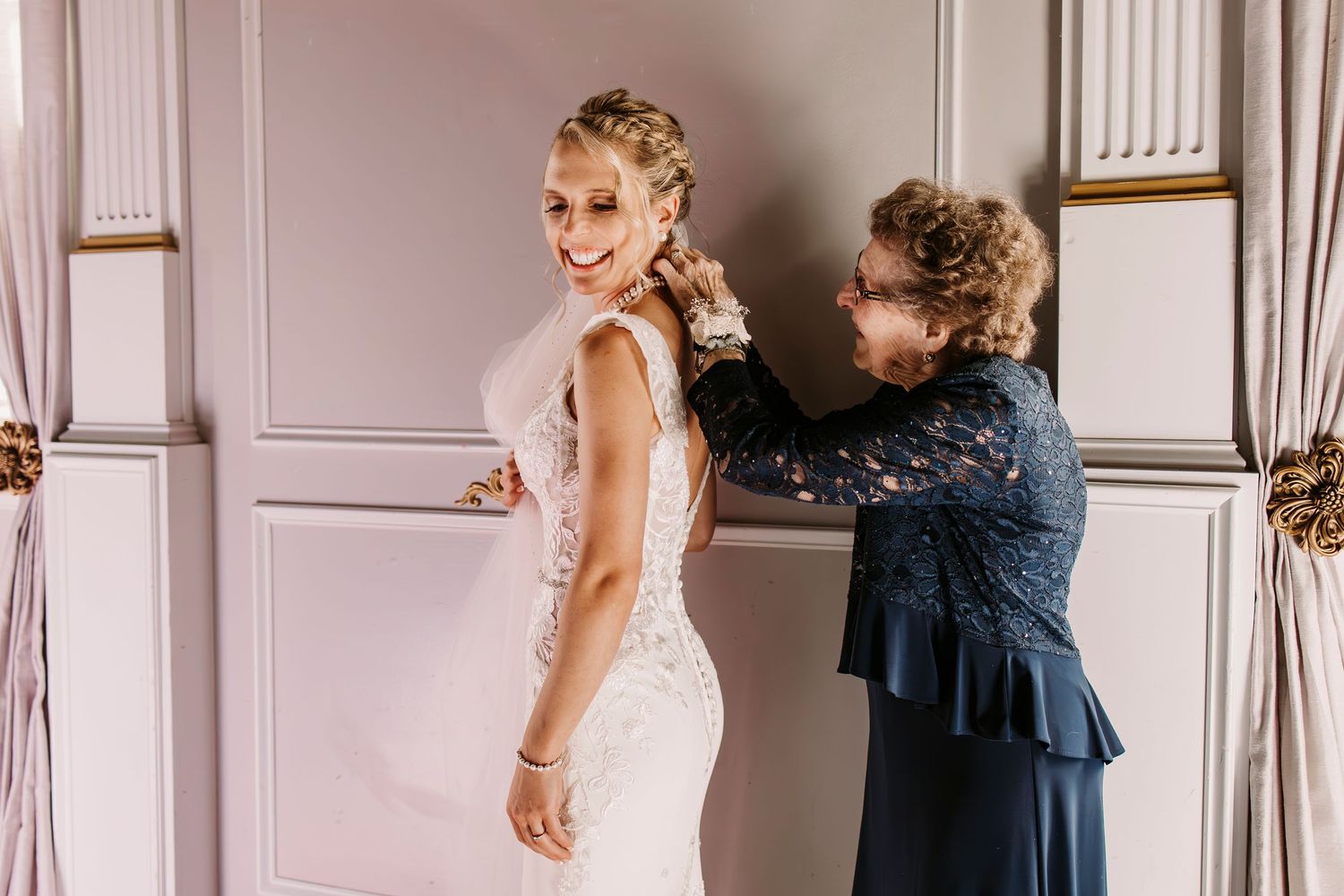 A bride in a fitted lace wedding dress gets help with final adjustments in an elegant room with white walls.