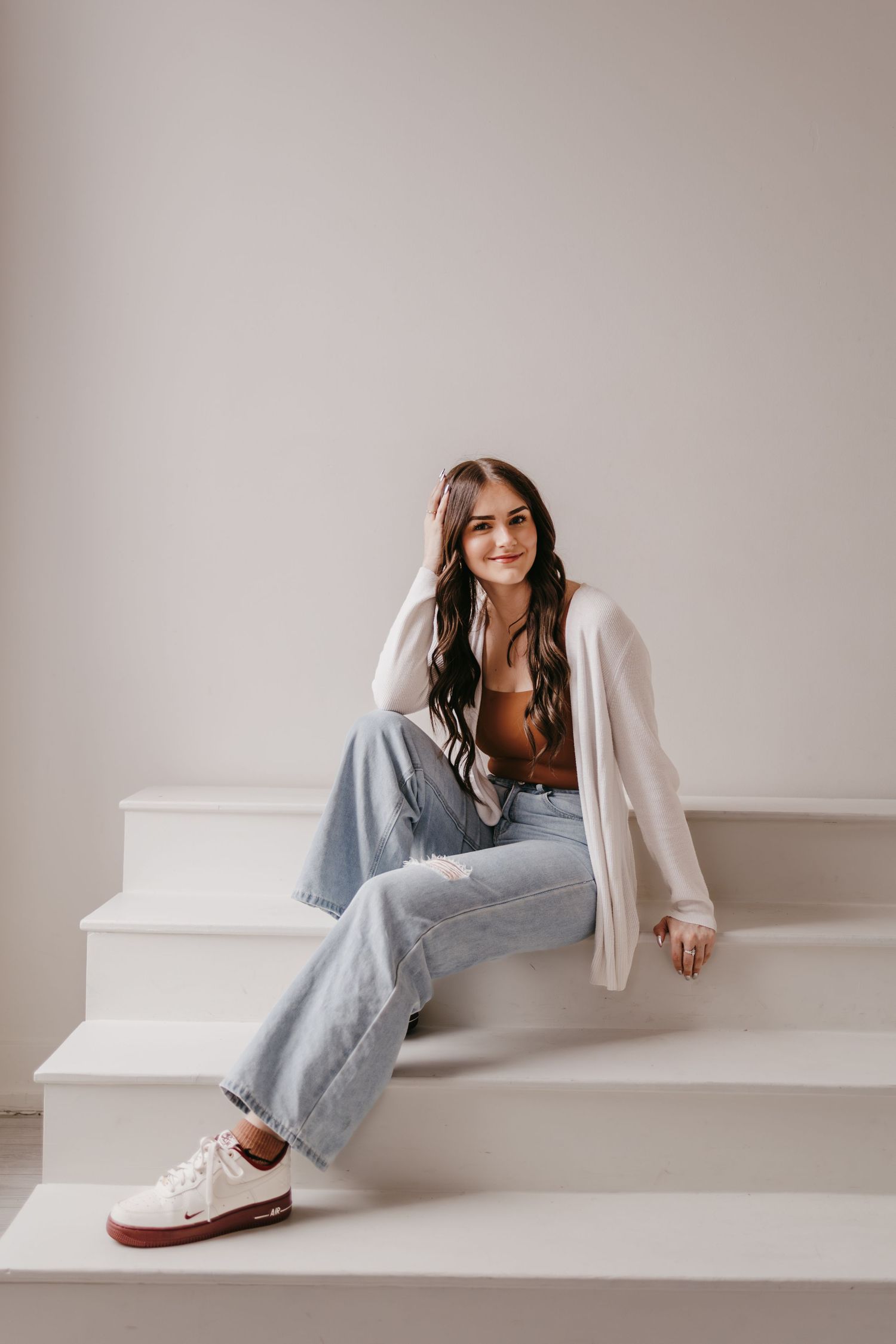 Woman in casual attire lounges against white wall wearing jeans and cardigan.
