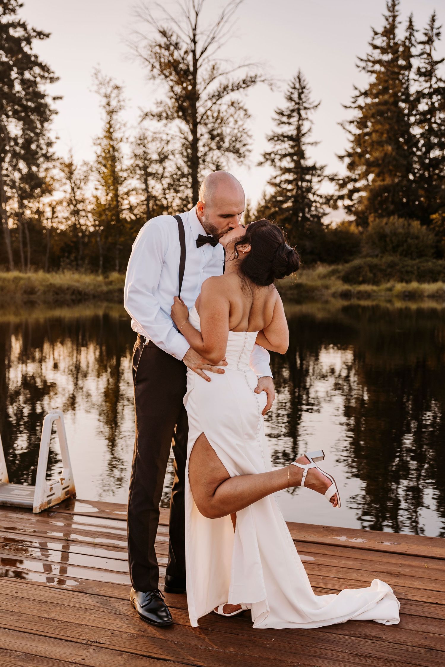 Newlyweds sharing an intimate moment on a wooden dock by a lake at sunset.