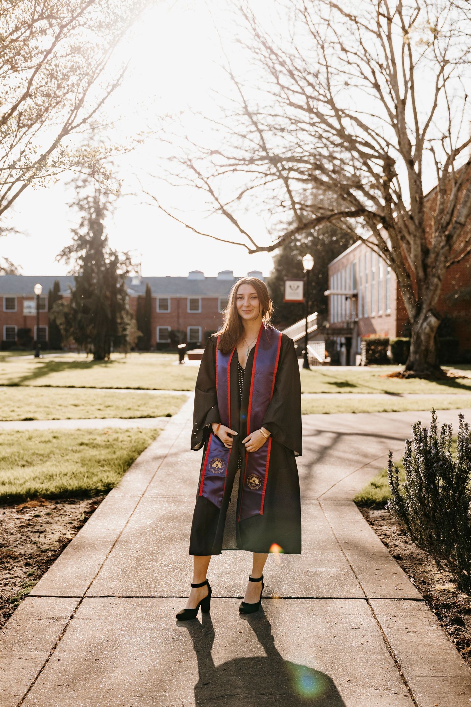Graduate in academic regalia stands on sidewalk during golden hour with brick buildings in background.