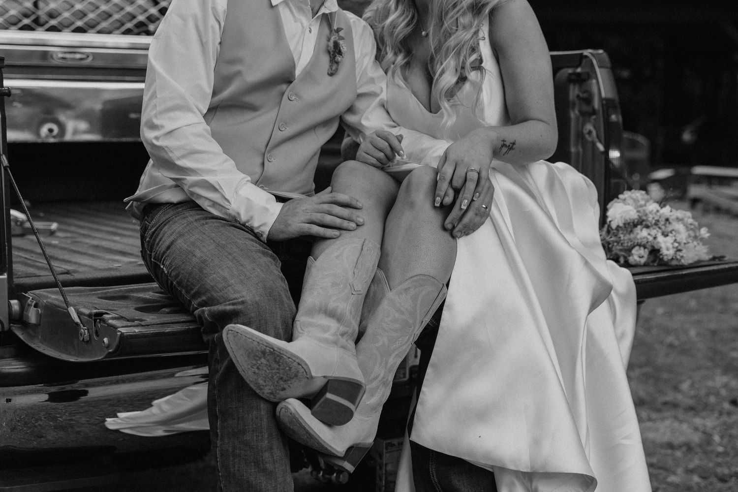Black and white photo of bride and groom sitting on truck tailgate, wearing cowboy boots and wedding attire.