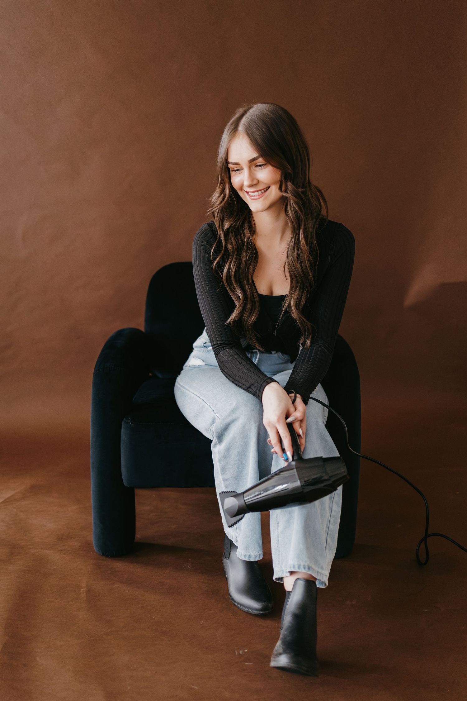 Model in black sweater and light jeans sits casually on dark chair against brown background.