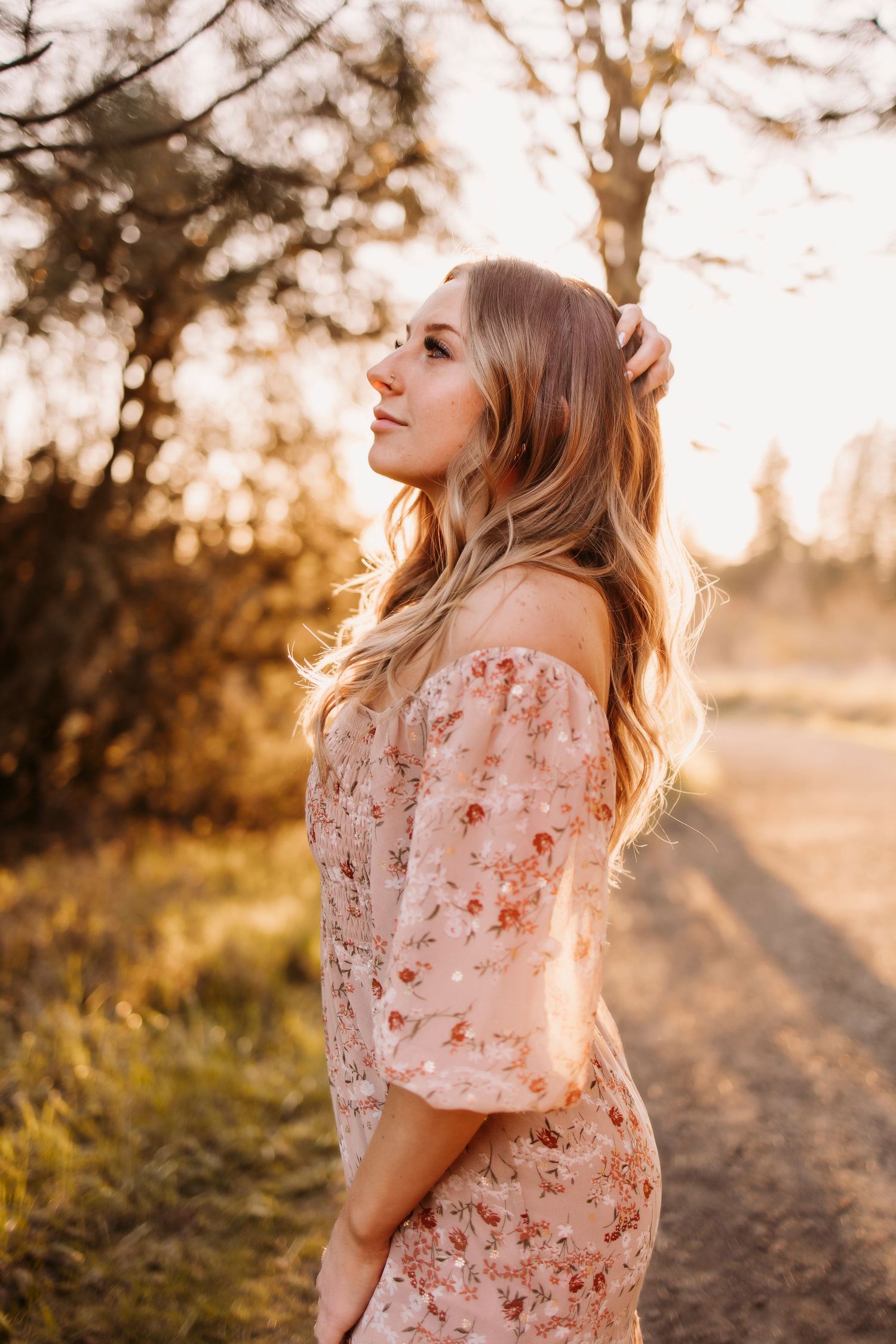 Woman in floral dress poses in golden sunset light on a dirt path surrounded by autumn foliage.