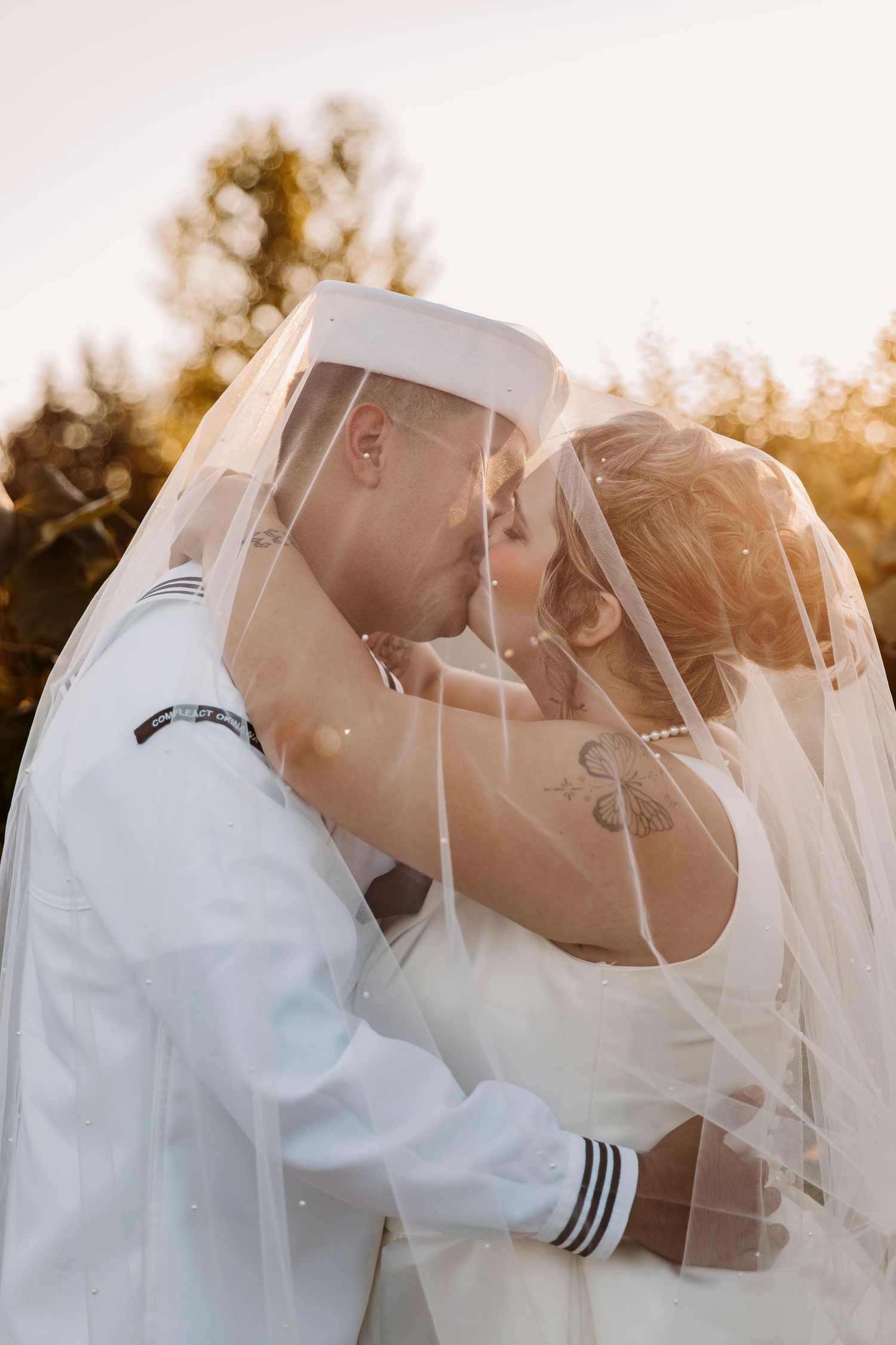 Naval wedding couple embraces under flowing veil at sunset.