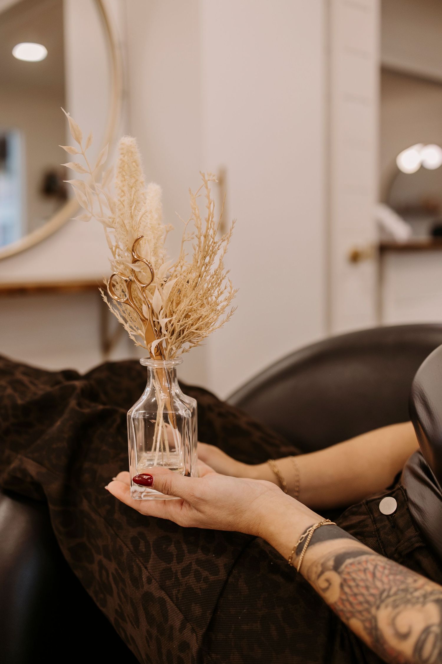 Tattooed hands holding small glass vases with dried flowers against a dark background.