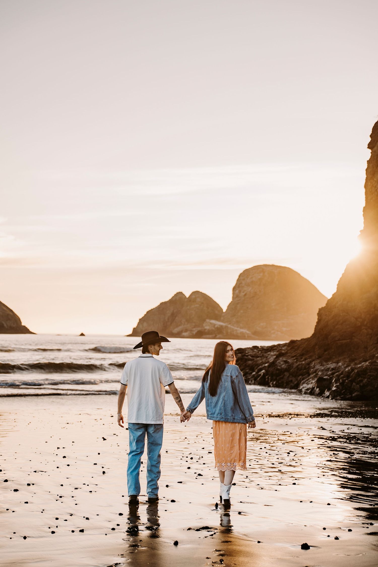 Couple walking hand in hand along a scenic beach at sunset with rocky cliffs in the background.