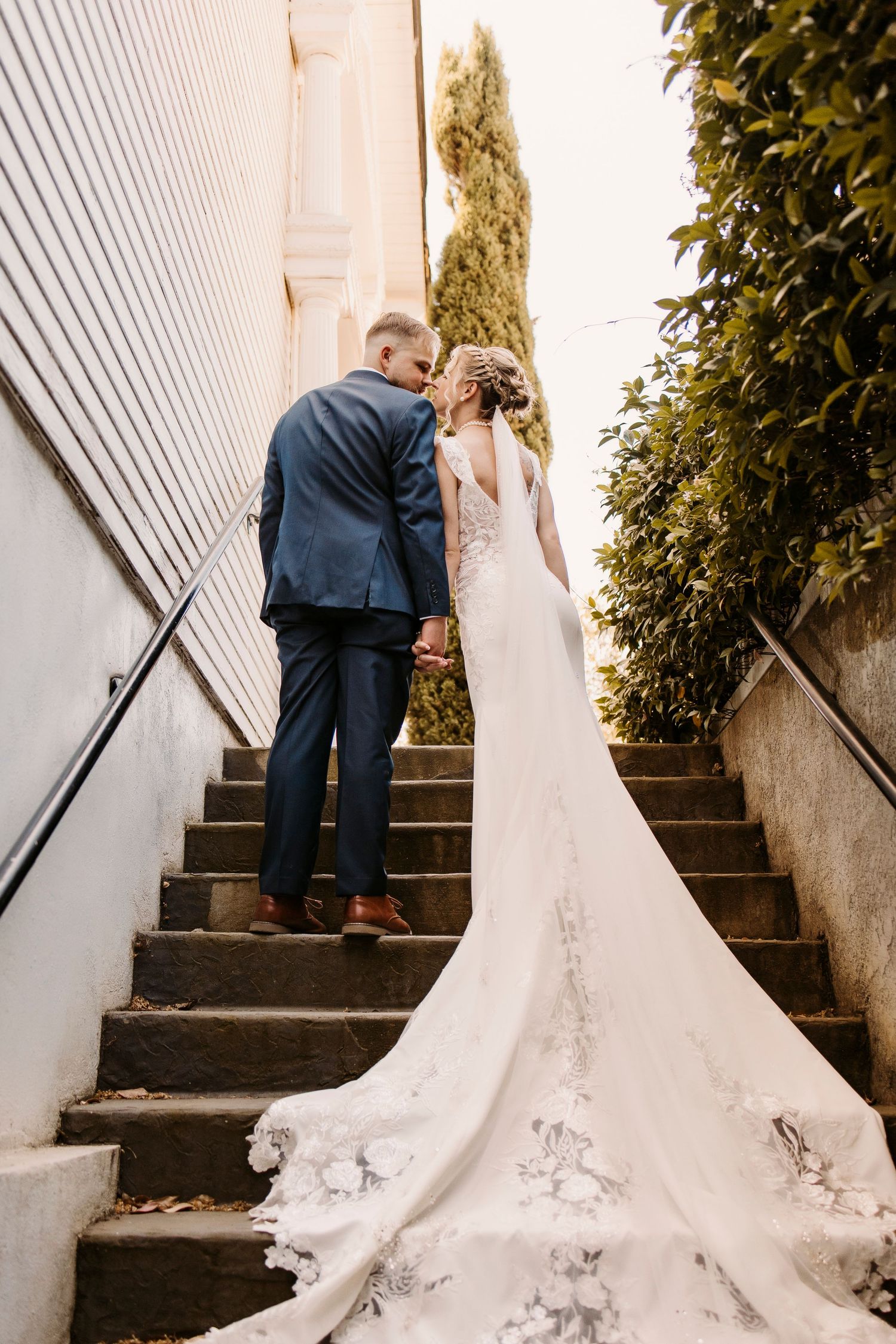 Wedding couple stands on outdoor steps with bride's long lace train cascading down the stairs.