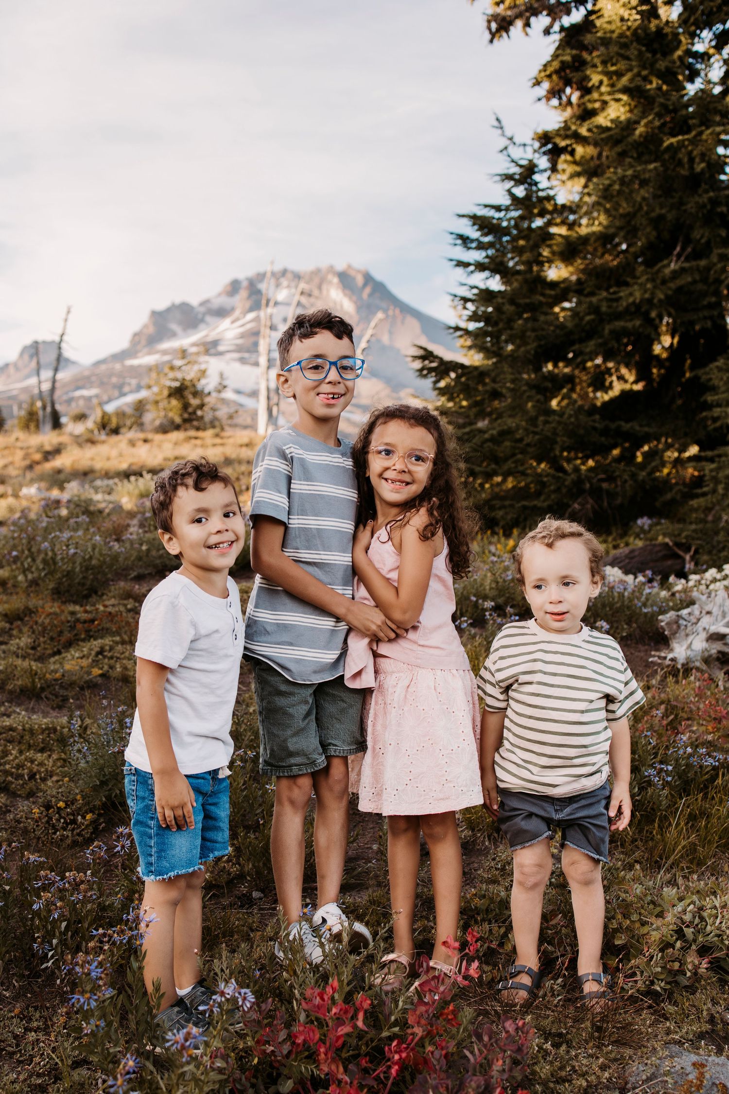Four children standing together on a mountainside with evergreen trees and peaks in background.