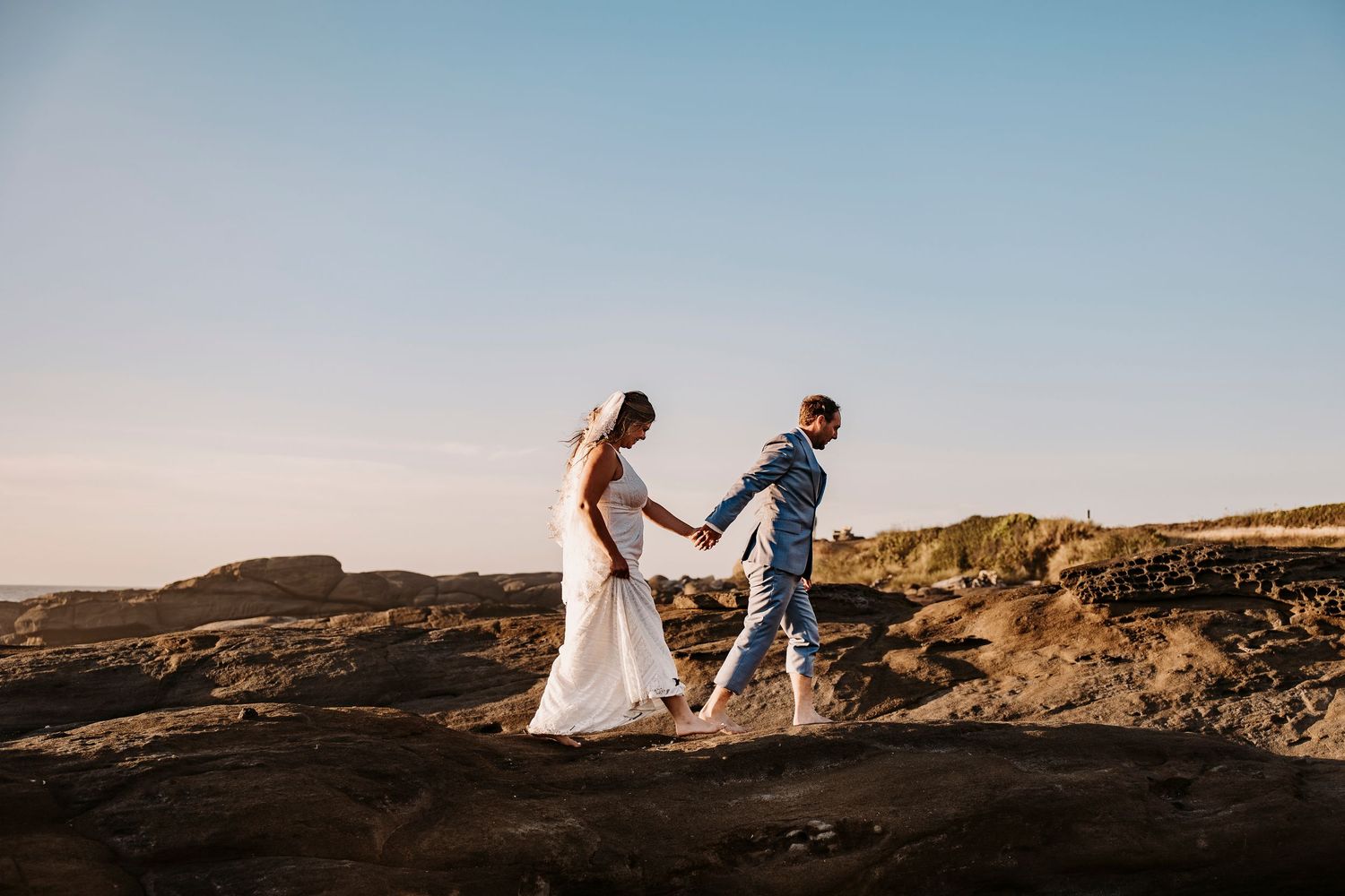 A couple in wedding attire walks together along scenic cliffs at sunset with ocean views in the background.